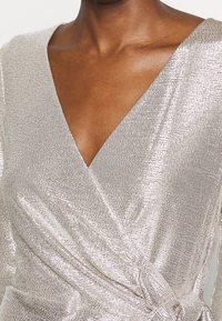 Metallisk wrapklänning i silver med djup V-ringning. Har en texturerad väv, mjuk drapering och en knytningsdetalj vid midjan.