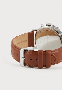 Bracelet de montre en cuir marron avec des coutures contrastées, boucle en métal argenté et partie d'un boîtier de montre en acier inoxydable visible.