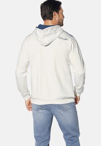White hoodie with a gray lining, adjustable drawstring, and embroidered logo on the back, paired with light blue jeans.