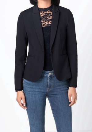 Femme portant un haut en dentelle noir sous un blazer noir ajusté, associé à un jean taille haute bleu moyen, debout avec les mains détendues.