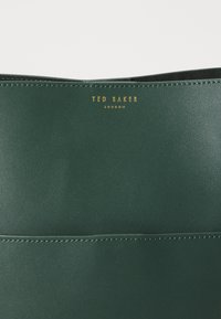 Superfície de pele verde escura com costura cuidadosa e logótipo dourado "Ted Baker London" embutido no centro superior.