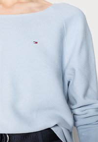 Pull tricoté bleu clair avec un large col, comportant un petit logo brodé sur la poitrine et des poignets côtelés. Texture sûre et douce.