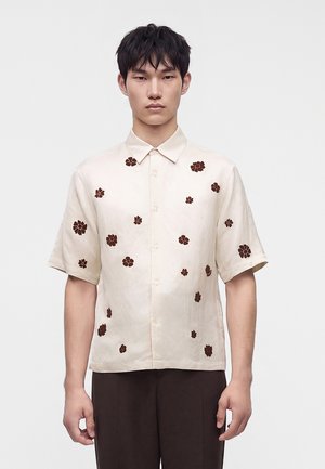 Jeune homme portant une chemise crème à manches courtes avec des motifs floraux marron, debout devant un fond clair uni.