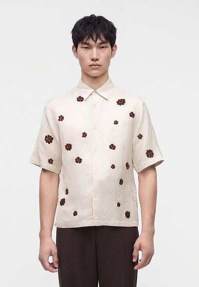 Jeune homme portant une chemise crème à manches courtes avec des motifs floraux marron, debout devant un fond clair uni.