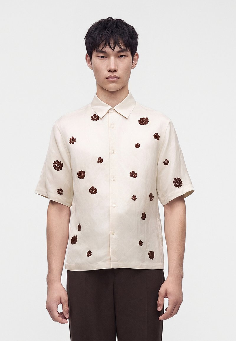 Jeune homme portant une chemise crème à manches courtes avec des motifs floraux marron, debout devant un fond clair uni.