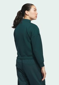Felpa verde con collo alto e polsini aderenti, caratterizzata da tre strisce verticali sulle spalle. Il materiale sembra morbido e texturizzato.