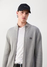 Cardigan grigio a coste con colletto alto, chiusura a zip e una patch con il logo. Indossato sopra una t-shirt bianca, completato da un berretto da baseball nero.