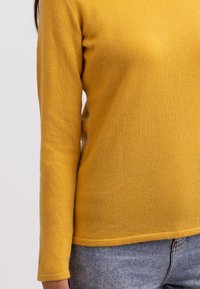 Gelber Strickpullover mit langen Ärmeln, rundem Ausschnitt und weicher Textur. Kombiniert mit hellblauen Jeans im schlichten Design.
