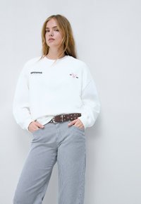 Sudadera de algodón blanca con logo y bordado de pájaro rosa. Combinada con pantalones a rayas grises y blancos y un cinturón marrón con tachuelas.