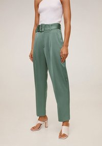 Pantalon vert à jambes larges en tissu lisse, avec une large ceinture et de nettes pinces à l'avant. Porté avec des talons blancs à brides.
