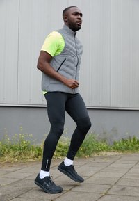 Athletic outfit featuring a gray quilted vest over a neon yellow shirt, paired with black leggings and black running shoes with white accents.