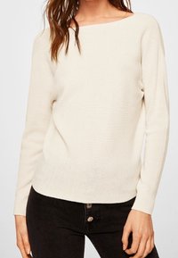 Cremefarbenes, gestricktes Sweatshirt mit weiter Ausschnitt, langen Ärmeln und einem strukturierten Muster. Kombiniert mit dunklen Jeans mit Knopfdetails.