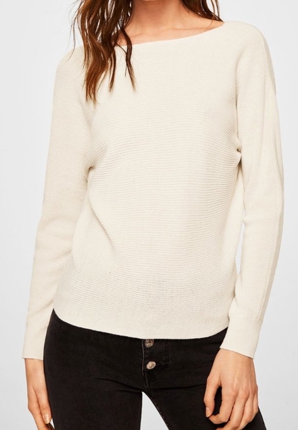 Cremefarbenes, gestricktes Sweatshirt mit weiter Ausschnitt, langen Ärmeln und einem strukturierten Muster. Kombiniert mit dunklen Jeans mit Knopfdetails.
