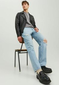 Chaqueta de moto de cuero negra sobre una camisa gris, combinada con jeans rasgados de un azul claro y mocasines negros, sentado en una silla de madera.