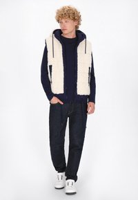 Vit fluffig väst över en marinblå hoodie, kombinerad med mörka jeans och vita sneakers. Vesten har två fickor och en dragsko längst ner.