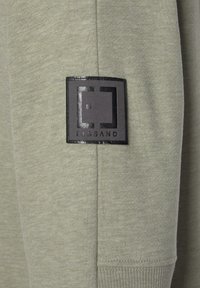 Tissu vert clair avec un patch carré noir présentant un logo géométrique stylisé et le texte "ELBSAND" en dessous.