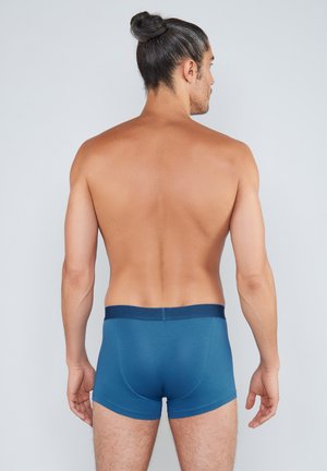 Hombre con el cabello largo recogido en un moño, vistiendo calzoncillos bóxer azules, de pie con la espalda hacia la cámara contra un fondo claro y liso.