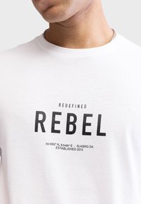 Hvid bomulds t-shirt med fed sort tekst: "REDEFINED REBEL" sammen med koordinater og "ESTABLISHED 2013" nederst.