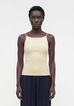 Γυναίκα με κοντά σγουρά μαλλιά, που φοράει μπεζ αμάνικο τοπ και φαρδιά μαύρη παντελόνια, στέκεται μπροστά σε απλή λευκή φόντο.