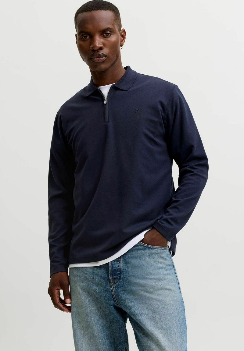 Homme noir portant un polo bleu marine à manches longues avec une fermeture éclair sur le devant et un jean bleu clair, debout avec une main dans la poche.