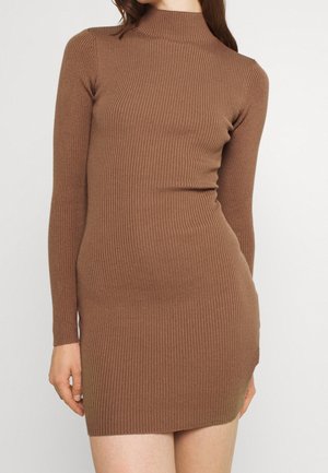 Femme portant une robe moulante en tricot côtelé marron à manches longues avec un col haut sur un fond clair uni.