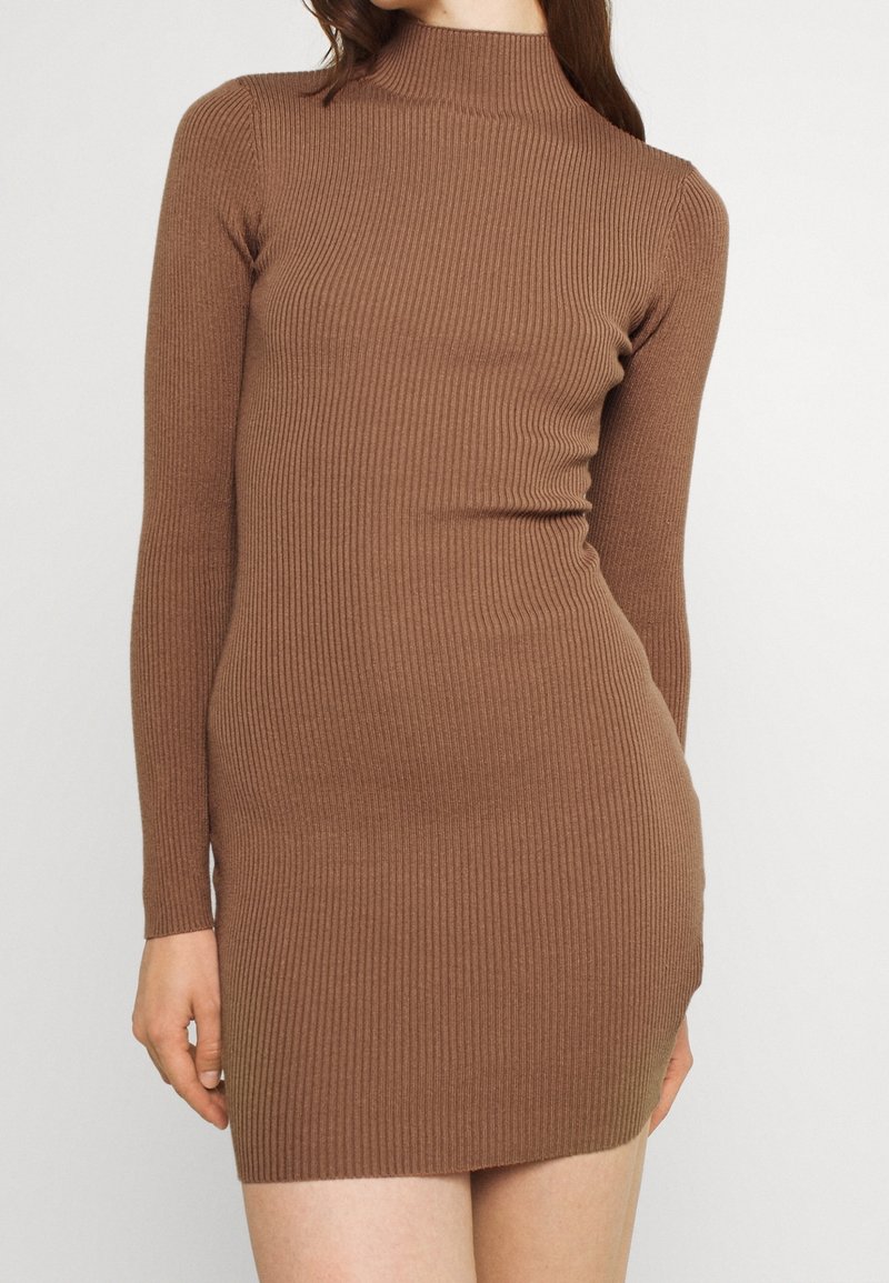 Kvinde iført en tætsiddende brun ribstrikket langærmet kjole med høj hals mod en ensfarvet lys baggrund.