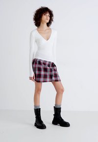 Weißes Langarmoberteil mit V-Ausschnitt, kombiniert mit einem karierten Rock in Rot, Schwarz und Grau. Das Model trägt schwarze Schnürstiefel mit grauen Socken.