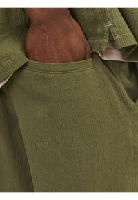 Khaki groene stof, licht gestructureerd met een gekreukt uiterlijk. Zak zichtbaar aan de zijkant, handpalm rustend binnenin, wat het ontwerp van het kledingstuk benadrukt.