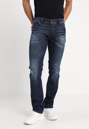 Mężczyzna w ciemnoniebieskich, dopasowanych jeansach z lekkimi przetarciami i białych trampkach, stojący na jednolitym jasnym tle.