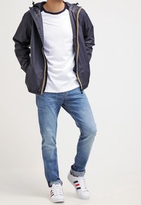 Marine let jakke med gul lynlås, lagdelt over en hvid T-shirt, parret med lyseblå jeans og hvide sneakers med røde og blå striber.