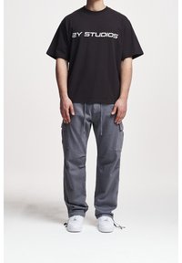 Osoba oblečená v černém tričku "2Y STUDIOS", šedých cargo kalhotách se šňůrkami a bílých teniskách, stojící před čistým bílým pozadím.