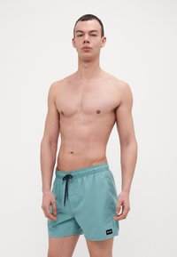 Světlomodré plavky vyrobené z hladkého, lehkého materiálu. Mají elastický pas se šňůrkou a malou nášivku s logem.