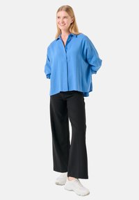 Chemise boutonnée oversize bleu clair avec col, portée sur un pantalon large noir et des baskets blanches. Tissu lisse avec une coupe décontractée.