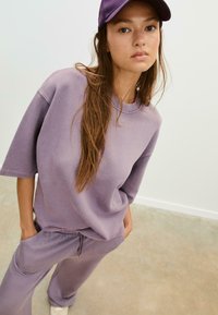 Fioletowy bawełniany oversize'owy t-shirt oraz pasujące spodnie, charakteryzujące się luźnym krojem i elastycznym pasem ze sznurkiem, w połączeniu z fioletową czapką baseballową.