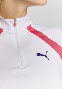 Bílý sportovní top se zipovým límcem, s růžovými akcenty na ramenech a tmavě modrým logem Puma na přední straně. Hladká, elastická tkanina.