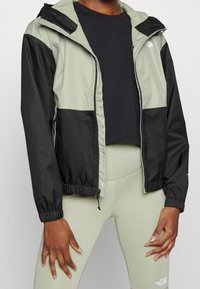 Jasnoszara i czarna kurtka przeciwdeszczowa z kapturem, elastycznymi mankietami i pasem. Noszona na czarnym krótkim topie i jasgreen leggings.