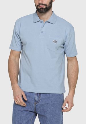 Uomo con polo azzurro manica corta con tasca frontale e piccolo logo ricamato, abbinata a jeans blu, in piedi davanti a uno sfondo neutro.