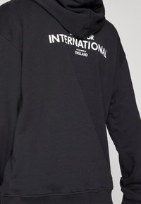 Černá mikina s přední klokaní kapsou, kapucí se stahovací šňůrkou a bílým nápisem „INTERNATIONAL“ na zádech. Jemná textura z bavlněné směsi.