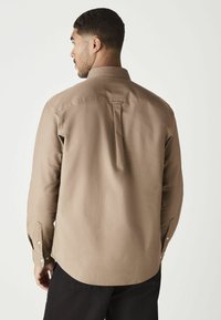 Hombre con cabello oscuro y corto lleva una camisa de manga larga beige y pantalones negros, de espaldas contra un fondo claro y liso.