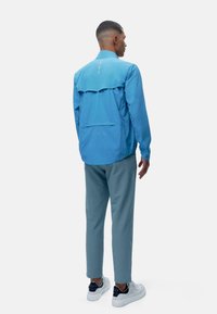 Kék, könnyű dzseki, hátul szellőző résszel, rugalmas mandzsettákkal és középső varrási részlettel. Világoskék nadrággal és fehér sportcipővel viselve.