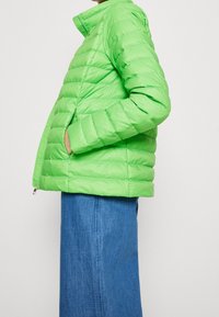 Doudoune vert clair avec motif matelassé horizontal, col montant, fermeture éclair et poches latérales, associée à une jupe en jean.