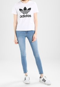 Camiseta blanca de algodón con logo negro de Adidas, combinada con jeans ajustados de color azul claro y zapatillas en blanco y negro. Diseño sencillo con un aspecto informal.