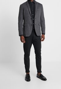 Blazer de lana gris con solapas de muesca, cierre de un botón y bolsillo en el pecho, combinado con una camisa negra y pantalones negros ajustados. Zapatos de cuero negros.