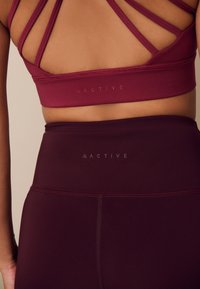 Reggiseno sportivo color melanzana con spalline incrociate e dettaglio logo, abbinato a leggings melanzana a vita alta con logo coordinato in vita.