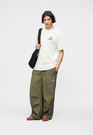 Persona joven con cabello hasta los hombros que lleva una camiseta blanca de Adidas, pantalones anchos color verde oliva, zapatos rojos y un bolso negro de hombro.