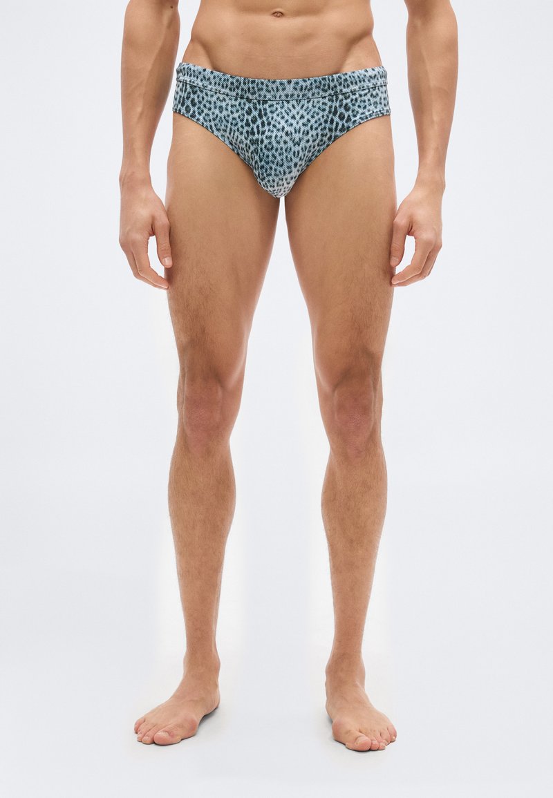 Άνδρας μοντέλο όρθιος, ξυπόλυτος, φοράει μπλε και μαύρα εσώρουχα με λεοπάρ σχέδιο, πάνω σε απλό φωτεινό φόντο.