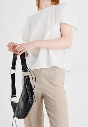 Woman wearing a white short-sleeve blouse and beige textured pants holding a black leather shoulder bag with braided details.