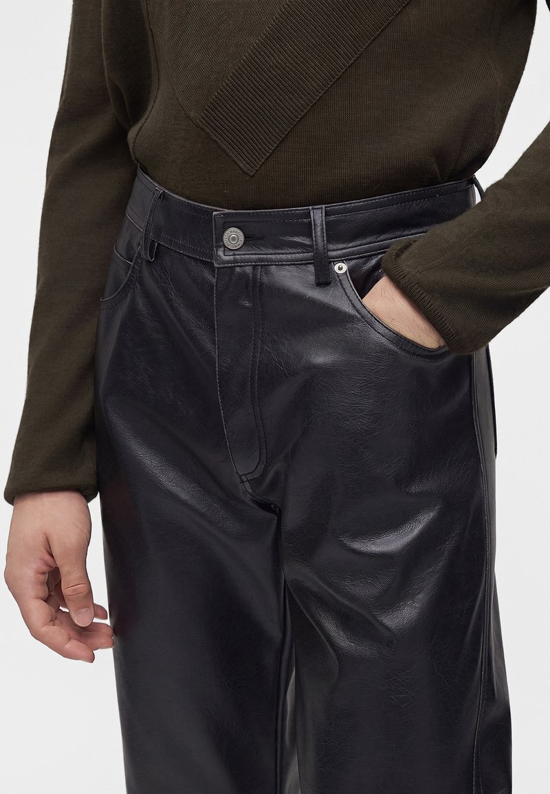 Person wearing black faux leather pants with a button and zipper, one hand in pocket, and dark olive knit sweater with ribbed detail.