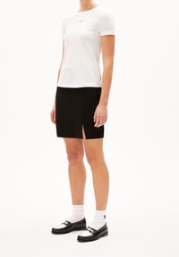 Žena ve bílém tričku s krátkým rukávem, černé sukni s předním rozparkem, bílých ponožkách a černobílých mokasínech na bílém pozadí.