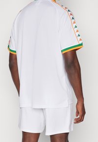 Bílý sportovní dres s oranžovými a zelenými akcenty, krátkými rukávy s vzorovaným lemem a volným střihem. V kombinaci s odpovídajícími šortkami.